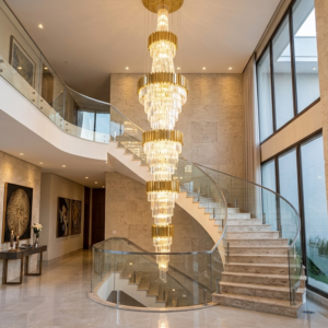 Fotografia de um hall de entrada residencial de alto padrão com pé-direito duplo. No centro do teto alto, um imponente chandelier moderno em camadas cascateia até a metade da altura, iluminando o espaço. Uma escadaria curva com degraus de mármore e guardas de vidro segue a curvatura da parede texturizada. O piso é de porcelanato polido e grandes janelas do chão ao teto dão para um jardim. Há um aparador com decoração no primeiro andar e um mezanino no segundo.