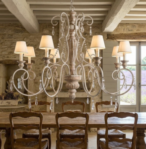 Lustre provençal de oito braços com acabamento envelhecido, cúpulas de tecido bege e cristais pendentes, centralizado em uma sala de jantar rústica com paredes de pedra e vista para campos de lavanda.
