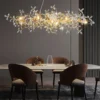 lustre-pendente-cristal-floral-moderno Lustre pendente horizontal de design orgânico com estrutura dourada e diversos cristais em formato de pétalas, iluminado sobre uma mesa de escritório e estante de livros cinza.
